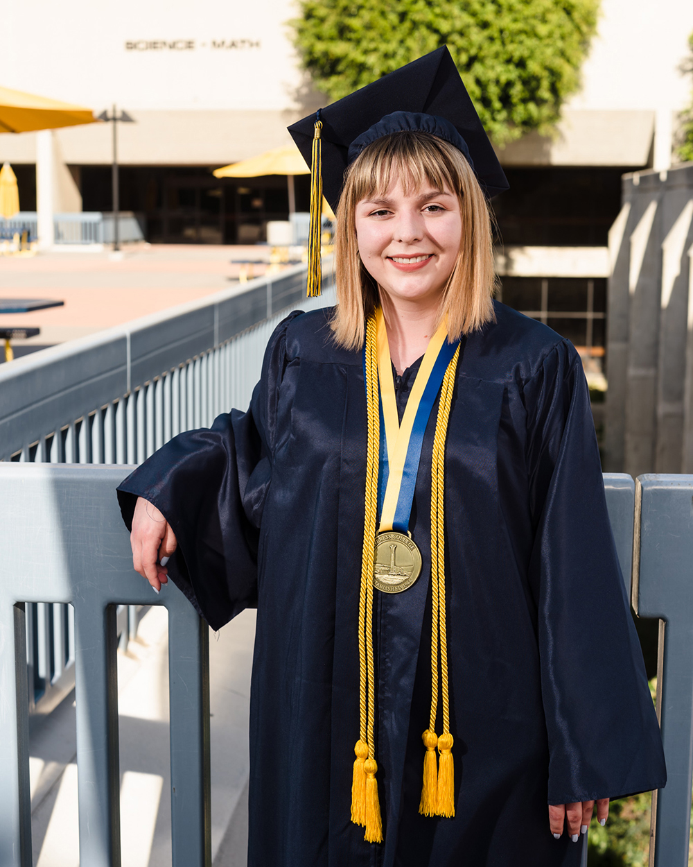 Student Bradi Palomarez stands in front of the Science and Math building on campus, wearing graduation regalia.