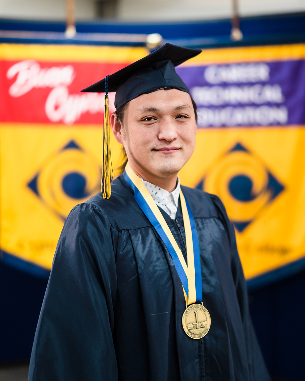 Culinary Arts student Dan Kim poses in graduation regalia in front of gonfalons for Cypress College and Career Technical Education