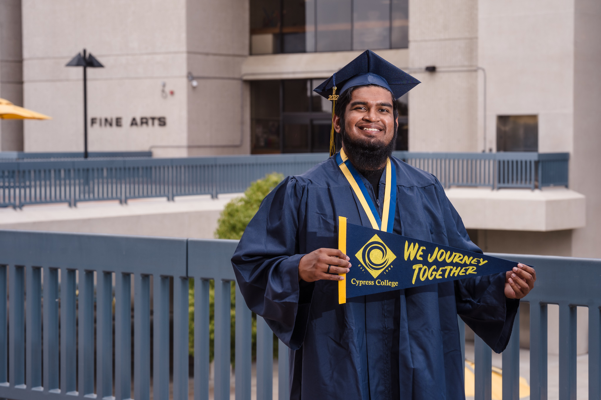 Student Abdul Meelar stands in graduation regalia in front of Gateway Plaza with green grass and buildings in background.