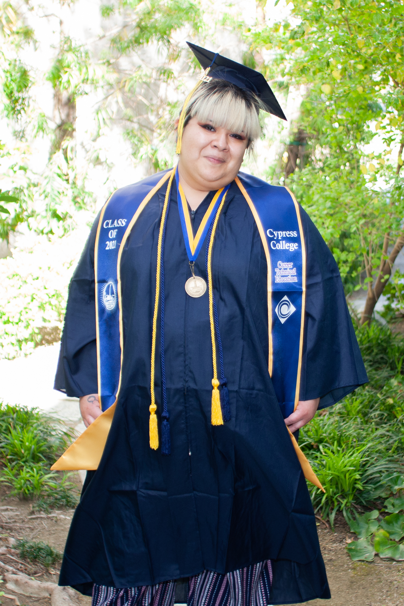 Alexandra Camacho poses beside trees on Cypress College campus wearing graduation regalia. 