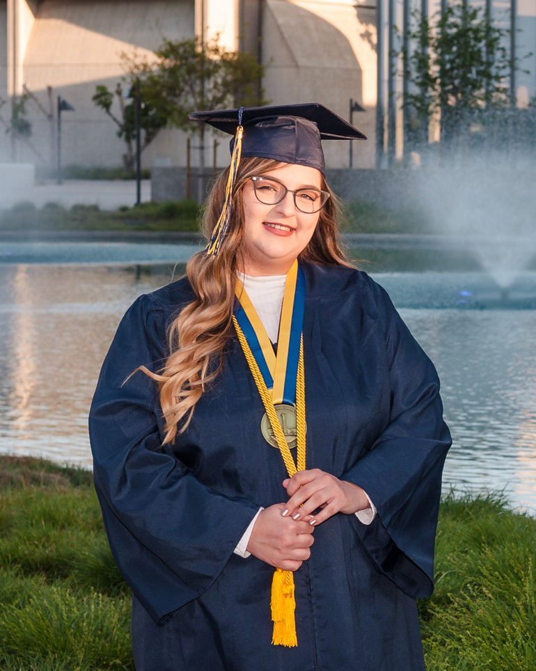 Student Krystal Kosack wears graduation regalia and poses in front of campus pond.