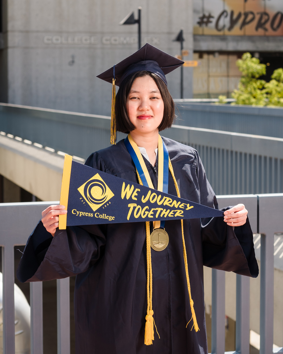 Student Pauline Lim poses with "We Journey Together" pennant as she wears graduation regalia and stands on the piazza.