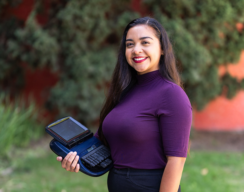 A woman with long hair and a purple shirt smiles as she holds a captioning machine on the campus of Cypress College.