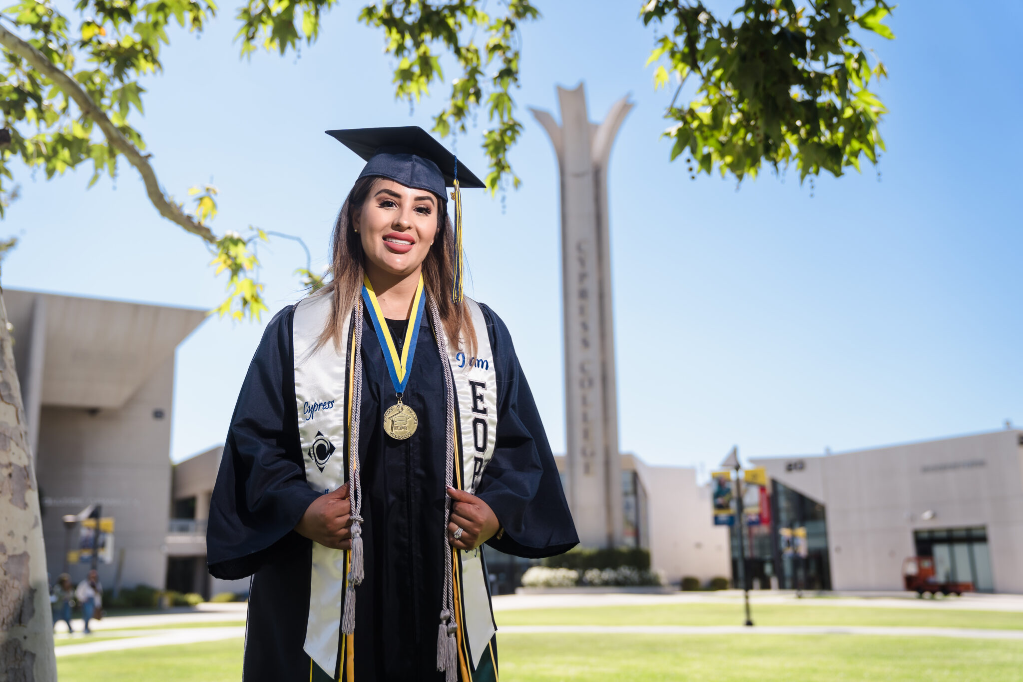 A smiling woman poses in college graduation regalia while posing in front of the Cypress College campanile. 