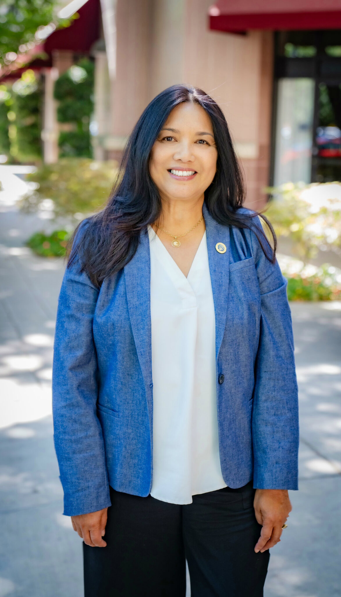 Woman with long dark straight hair stands outside with a blue jacket and white shirt