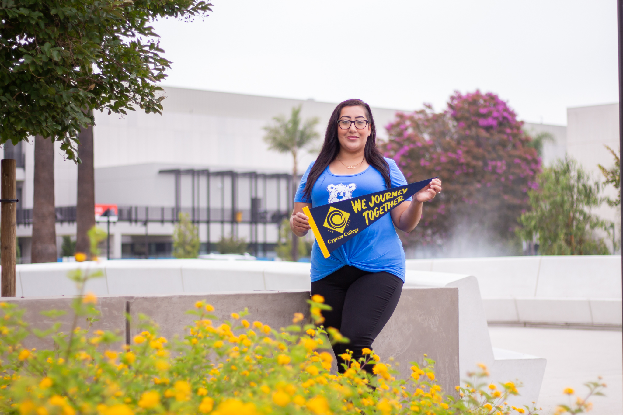 Student Melissa Whitewater stands in front of the Cypress College pond with the Science, Engineering and Math building, as she holds a "We journey together" pennant.