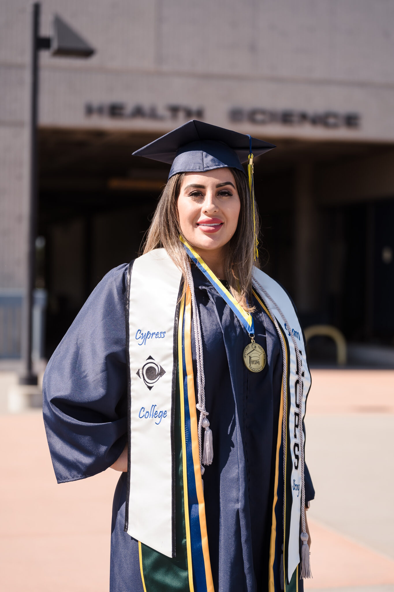 A woman in a graduation cap and gown stands in front of the Cypress College Health Science building. 
