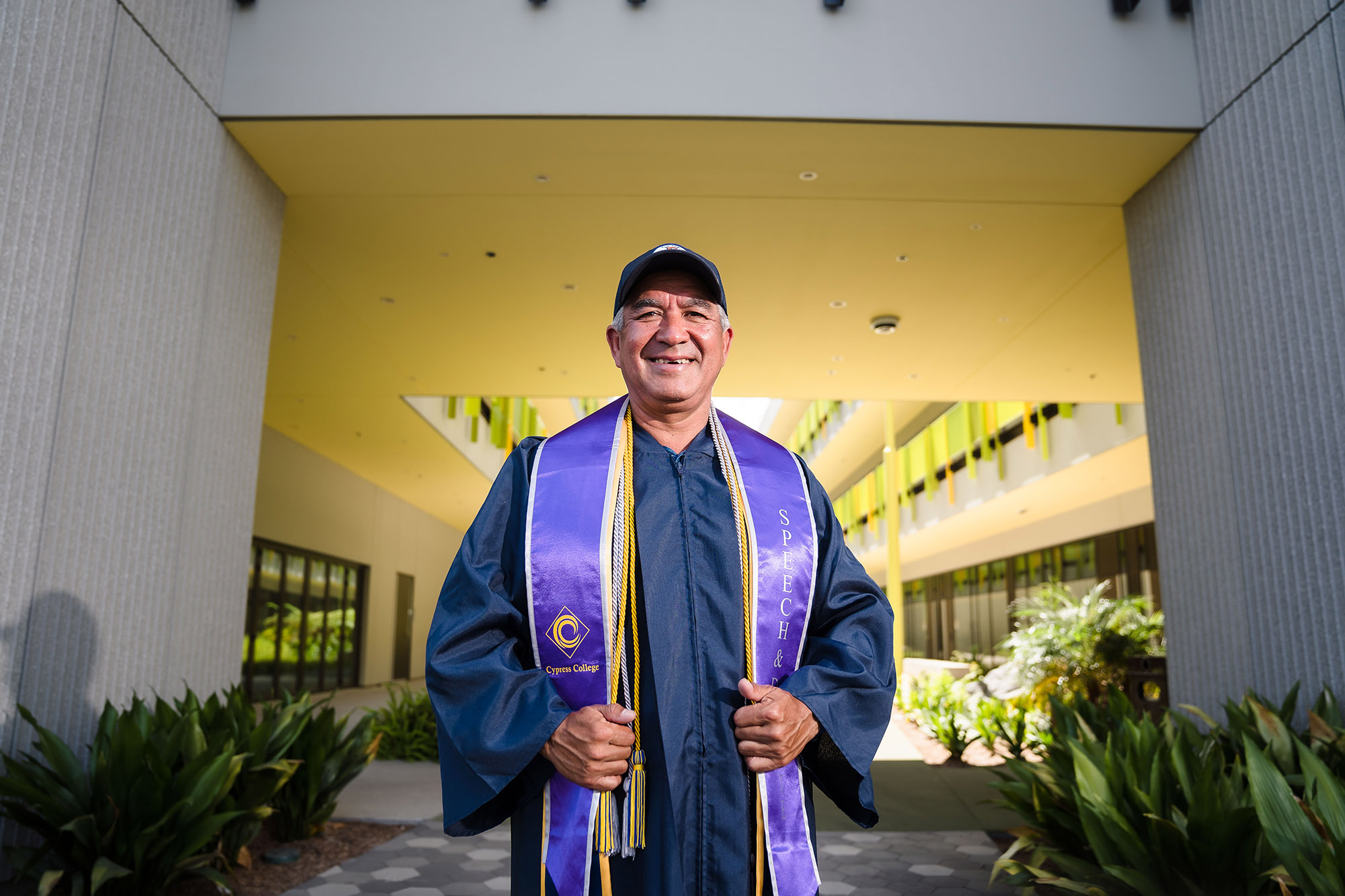 An older man in graduation regalia with honors cords and sashes poses in front of the Cypress College science building. 
