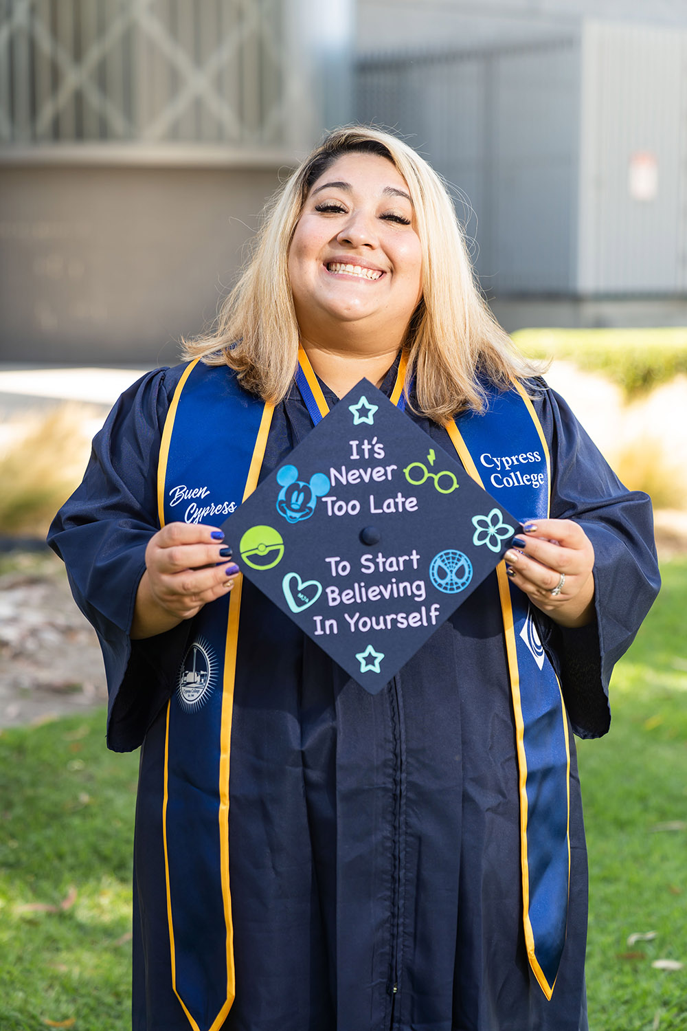 A woman in graduation regalia shines a bright smile as she holds up her mortarboard, which is decorated with the words: "It's never too late to start believing in yourself."