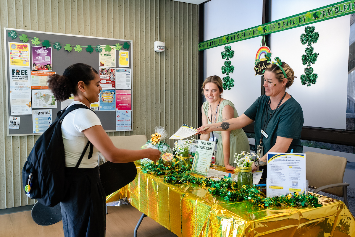 Two women in green at a table covered with St. Patrick's Day decorations give handouts to students.