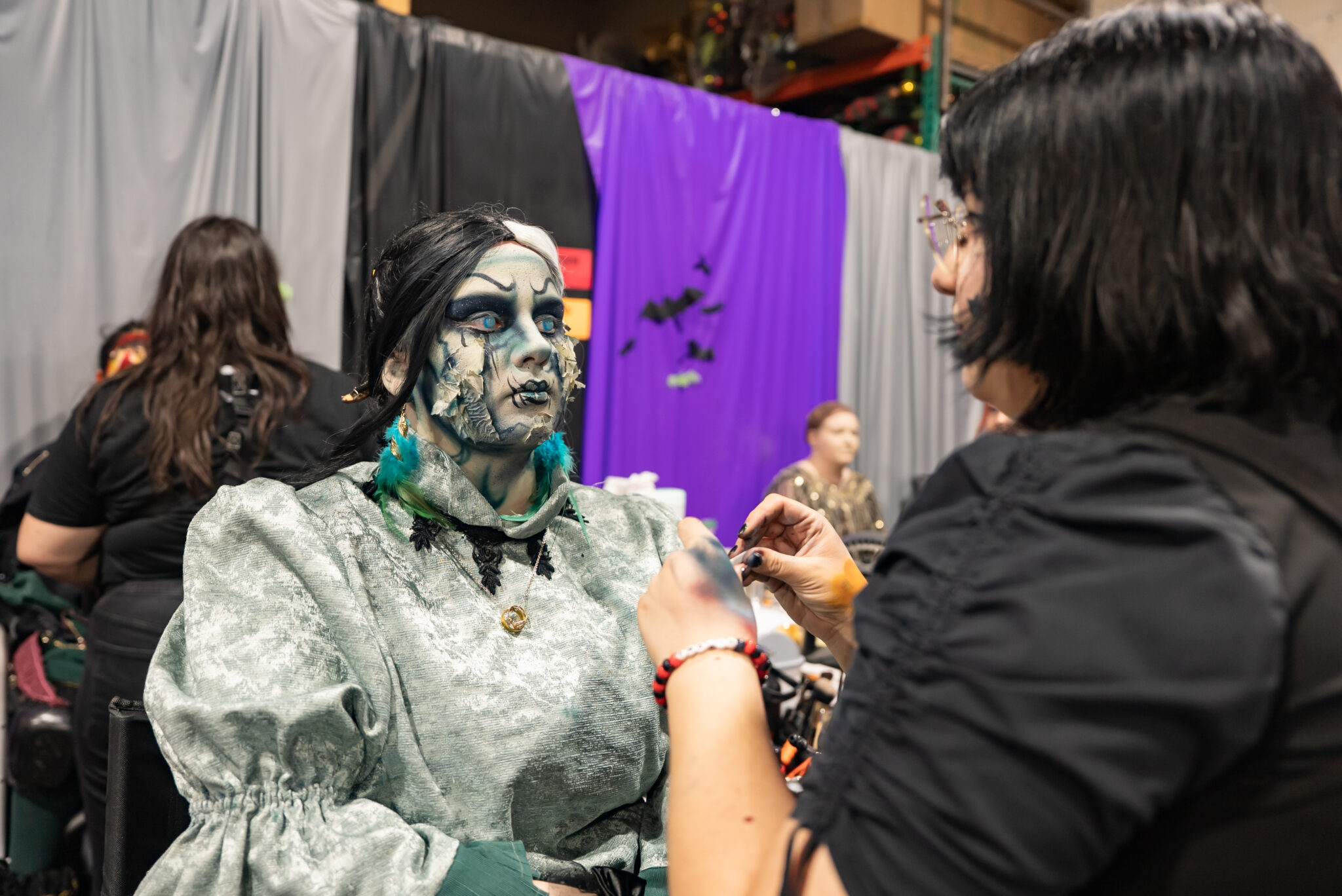 A scare actor wears a pale blue dress, long black wig, and waits for a makeup artist to apply pale blue/green makeup to her face, which has a ghostly quality.