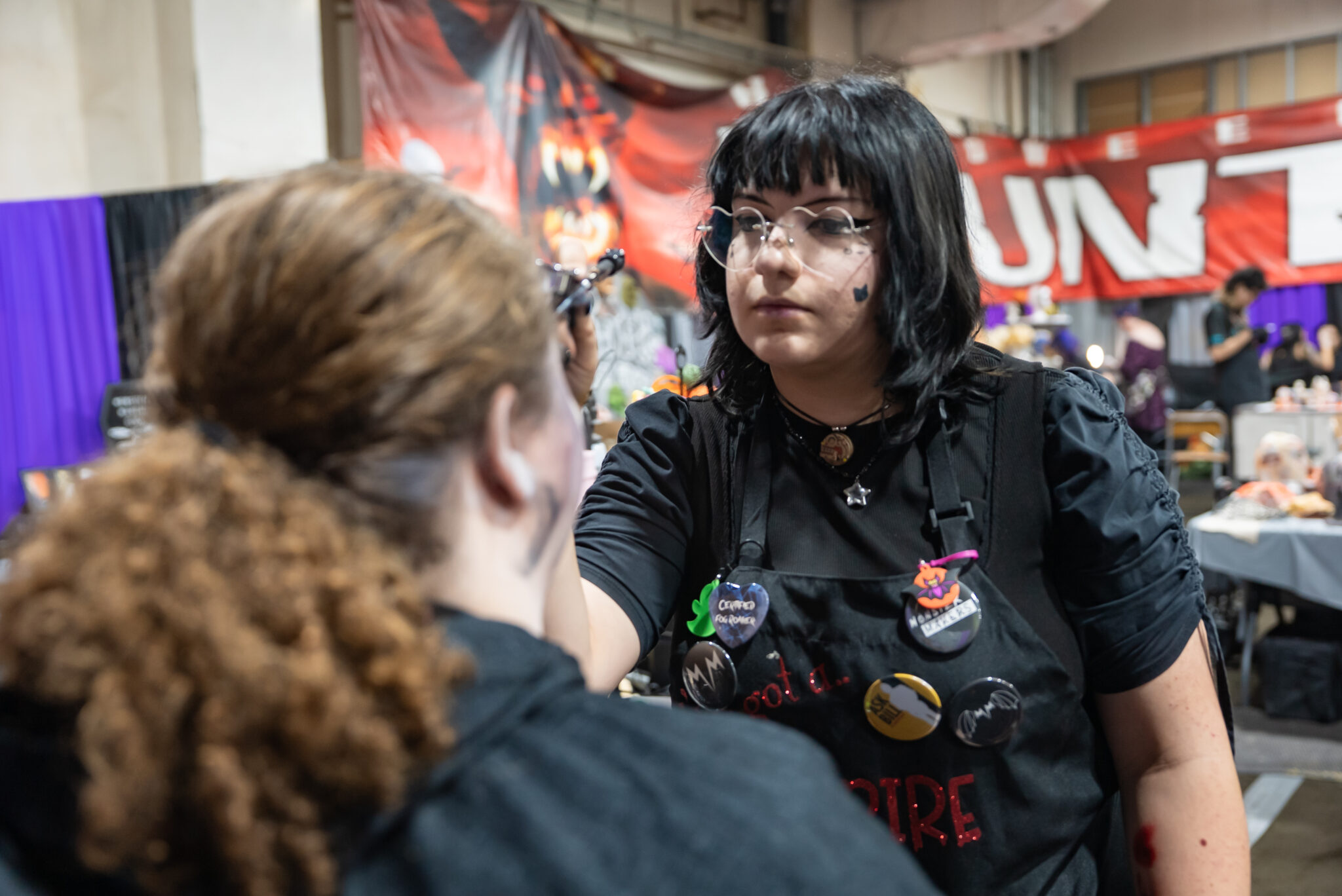 A woman facing the camera applies Halloween makeup to an actor