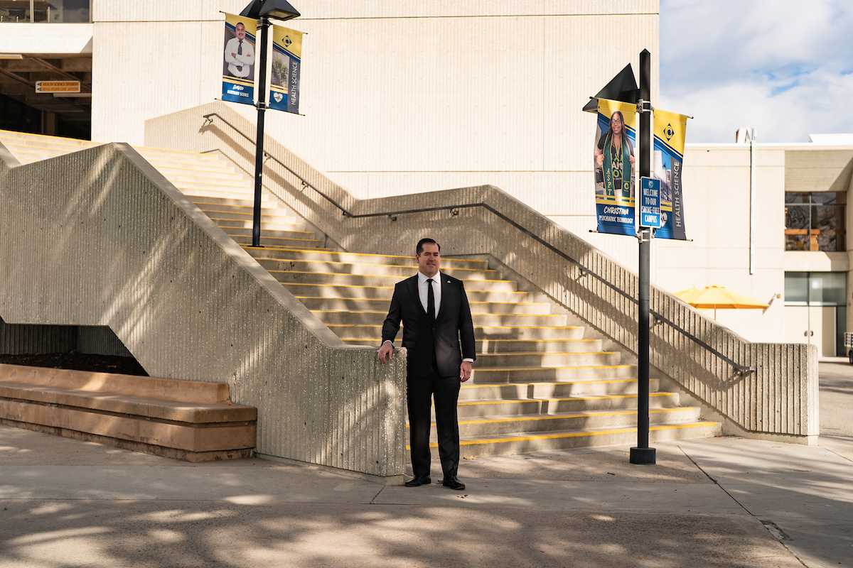 A man in a dark suit stands in front of a wide concrete staircase that leads to Health Science classes.