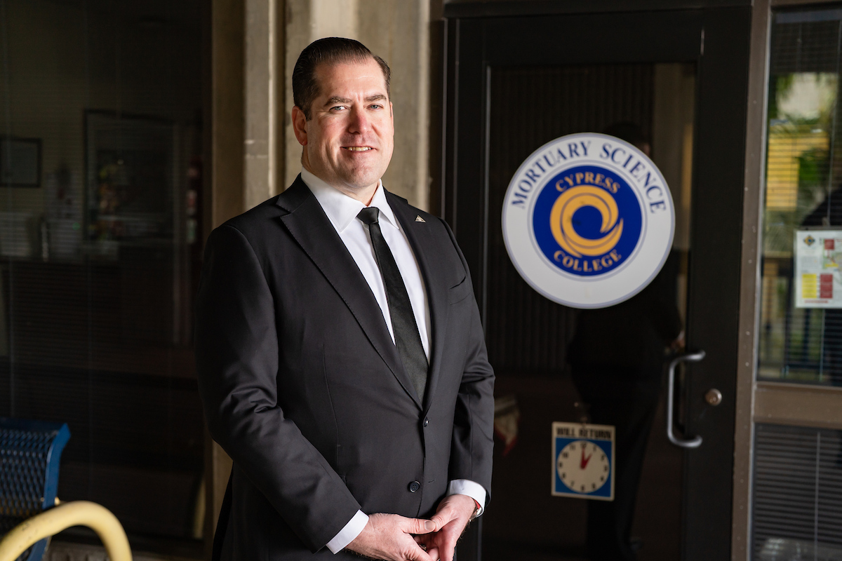 A man in a dark suit and tie stands in front of the Cypress College Mortuary Science Department door, identified by the logo on the glass.