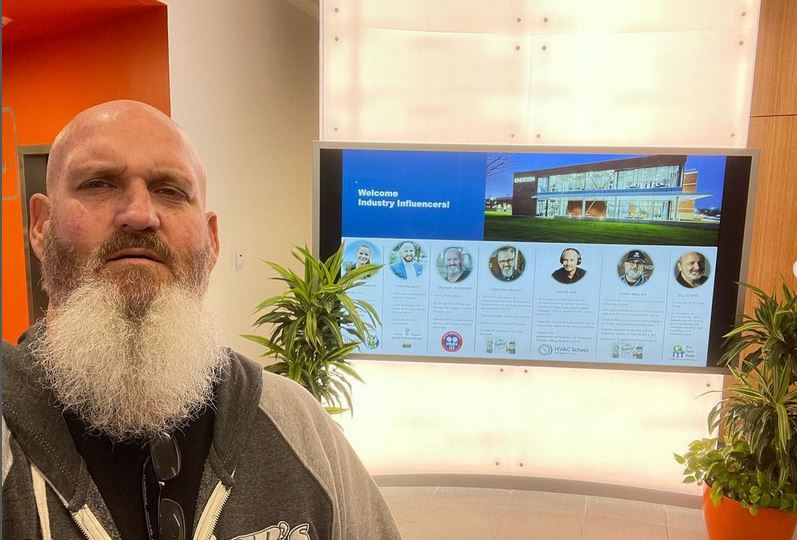 A bearded man stands in front of an event slide announcing that he is one of the key speakers at an event. 