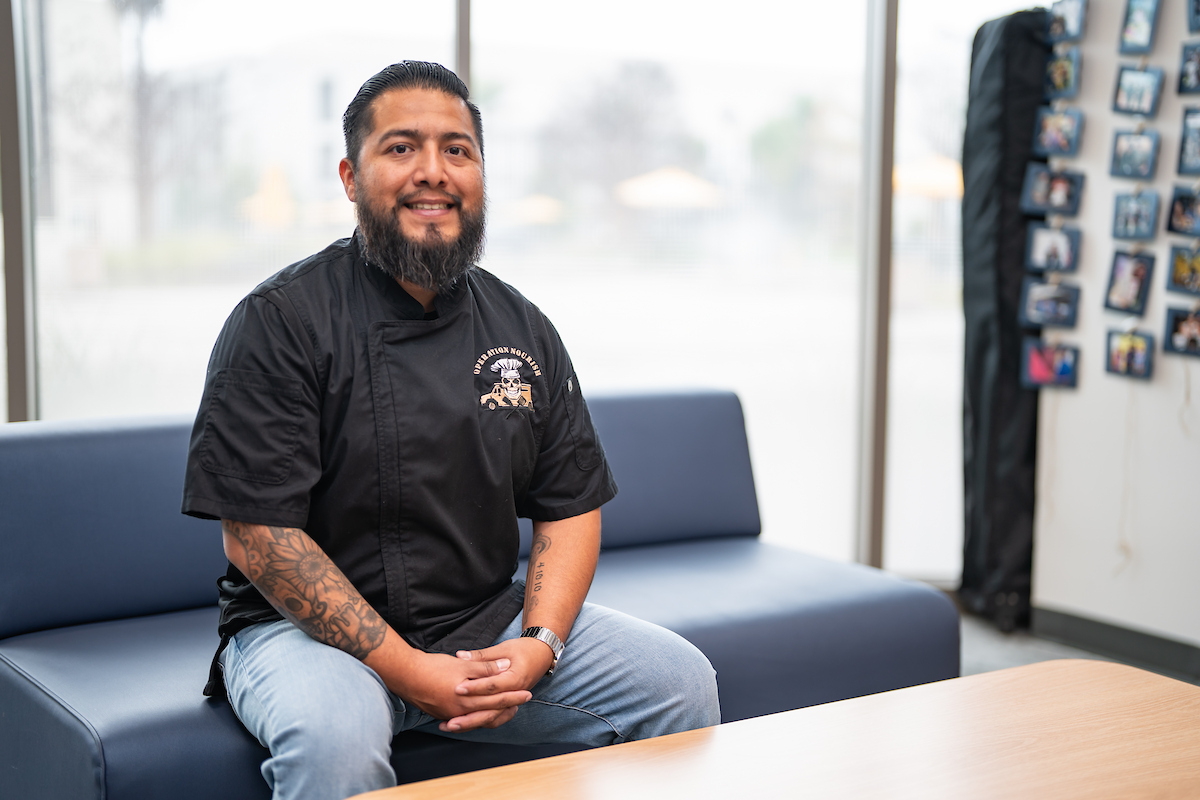 A man with black hair and beard smiles and sits in front of a window.