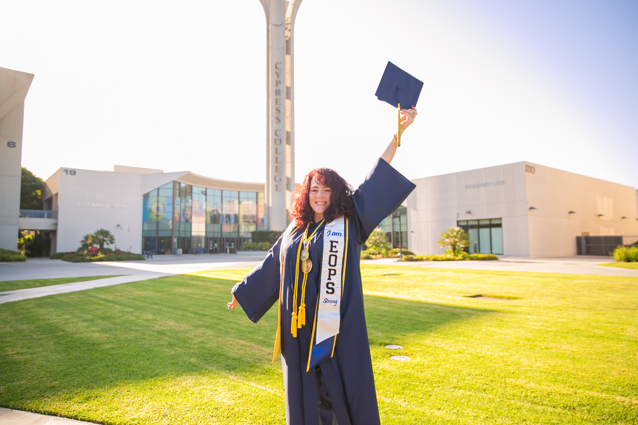 Woman holds graduation cap high in front of Cypress College campanile.