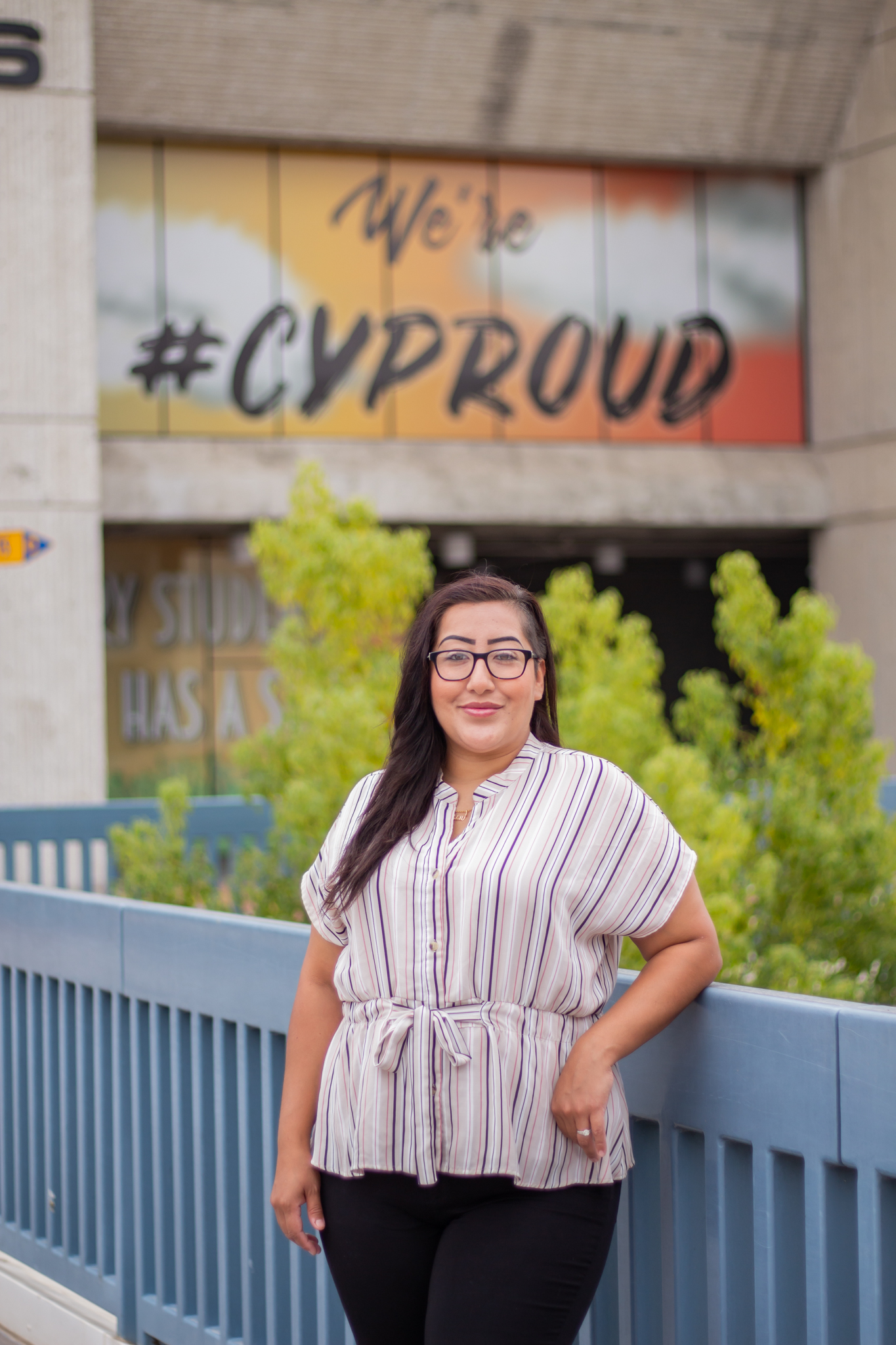 Cypress College student Melissa Whitewater wears a casual shirt as she stands on the second-level piazza on campus in front of a CYProud sign.