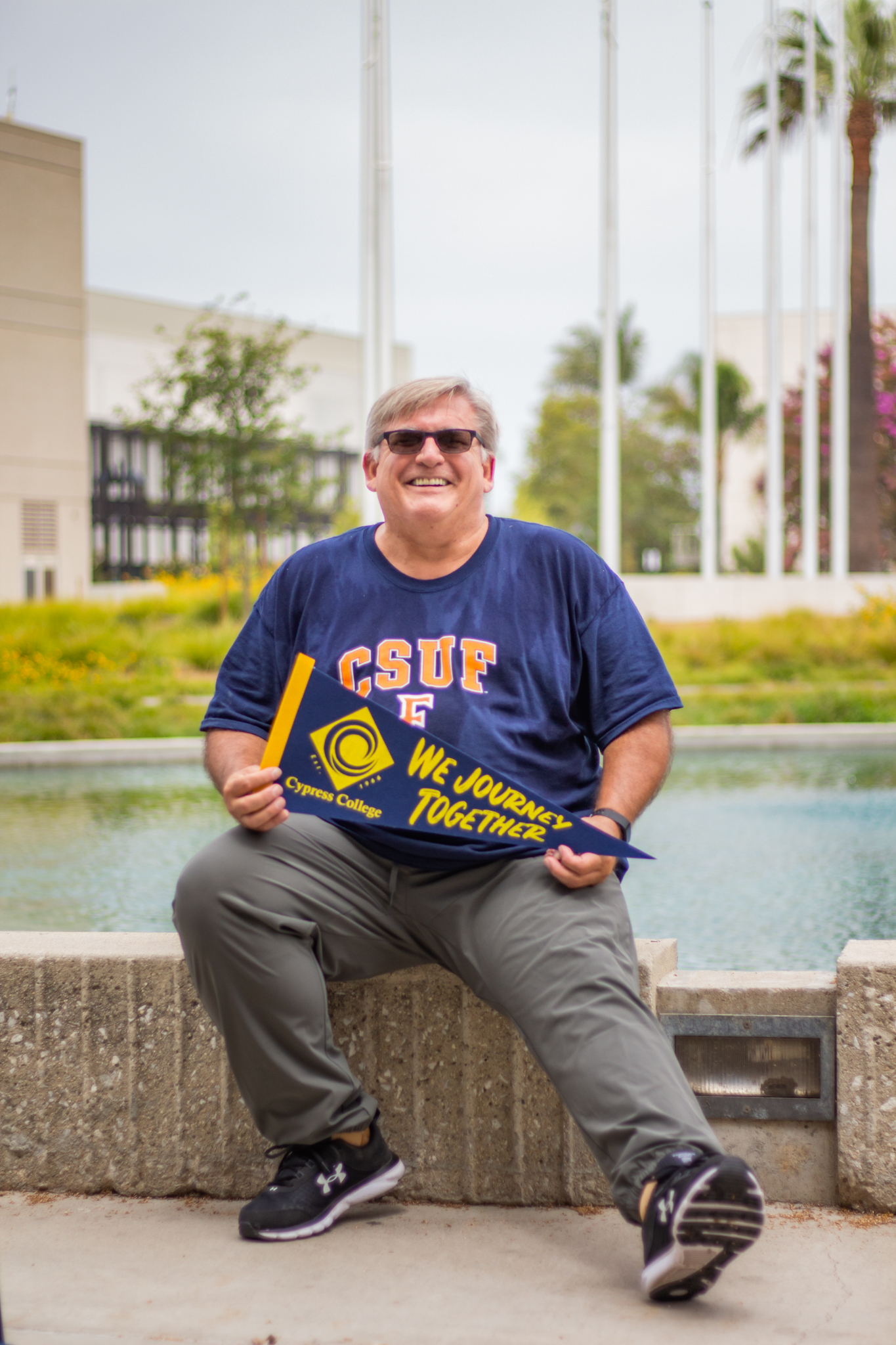 Patrick Hale sits at the edge of the Cypress College pond, holding a college pennant as he smiles at the camera.