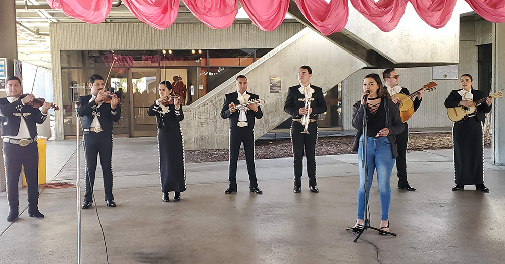 A mariachi band performs outdoors under a red banner. 