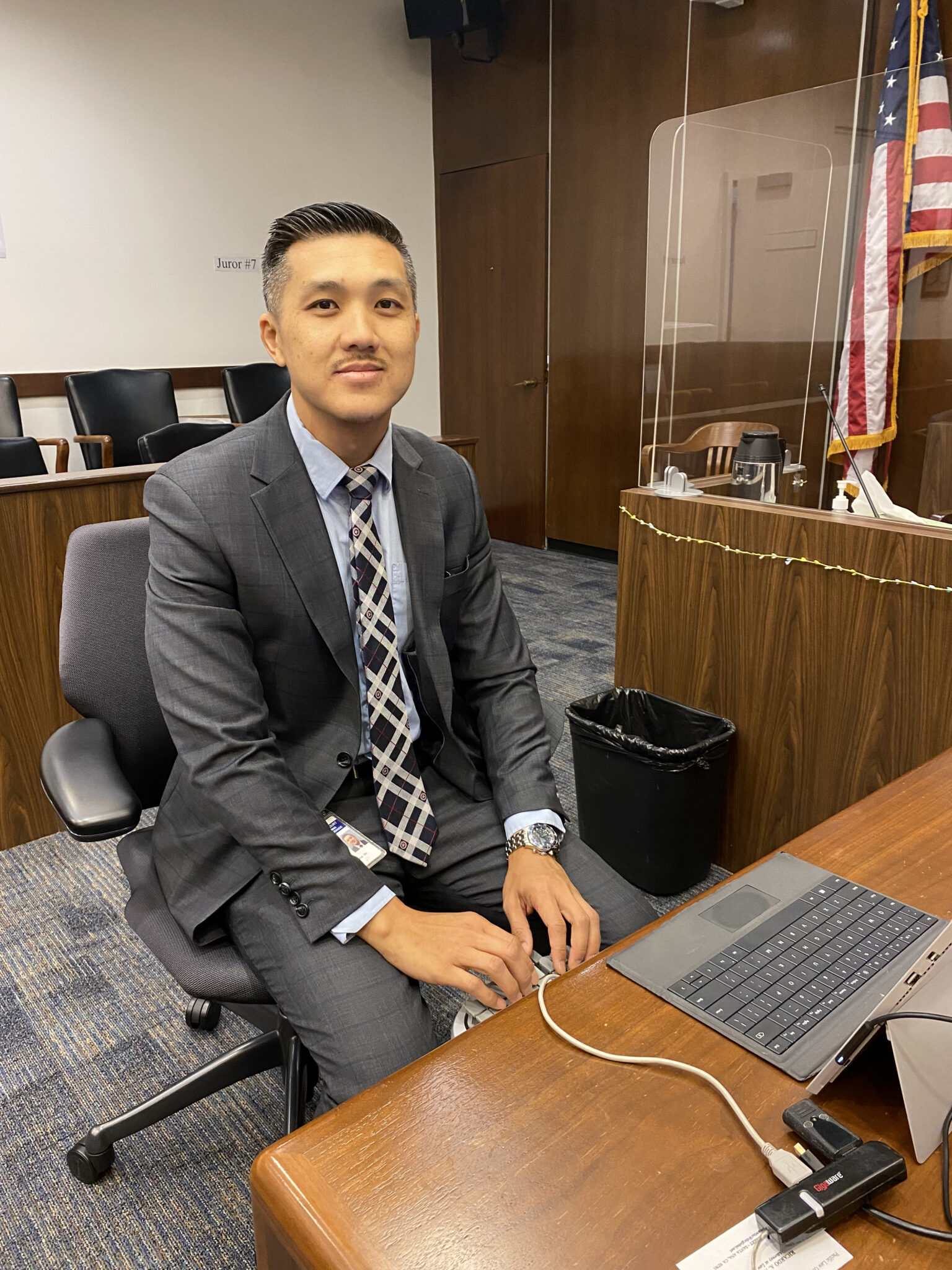 A man wearing a suit and tie sits at a keyboard and shorthand dictation machine in a courtroom.