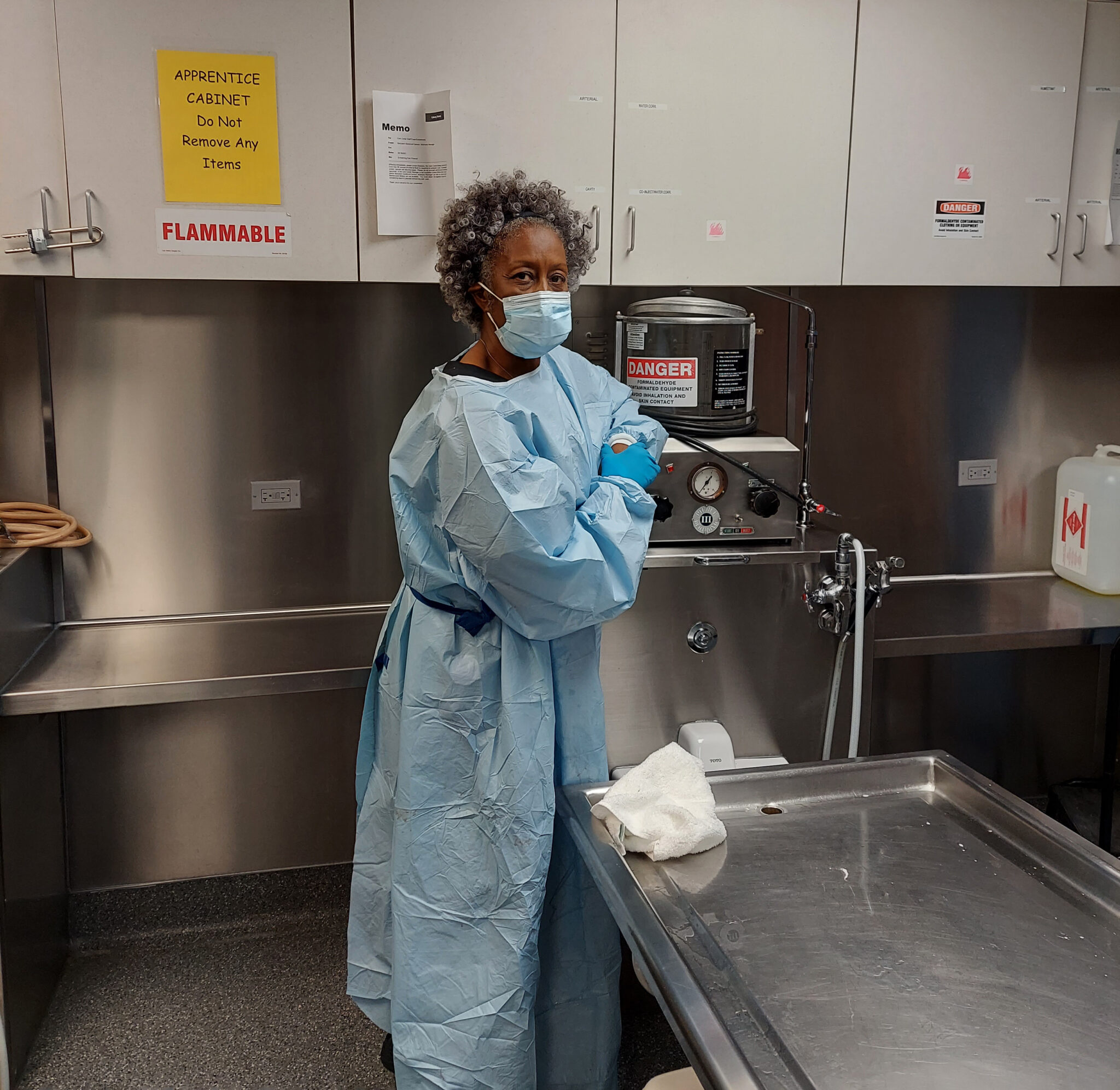 A woman with short curly hair, Margo Malone, wears medical protective gear at an embalming station. 
