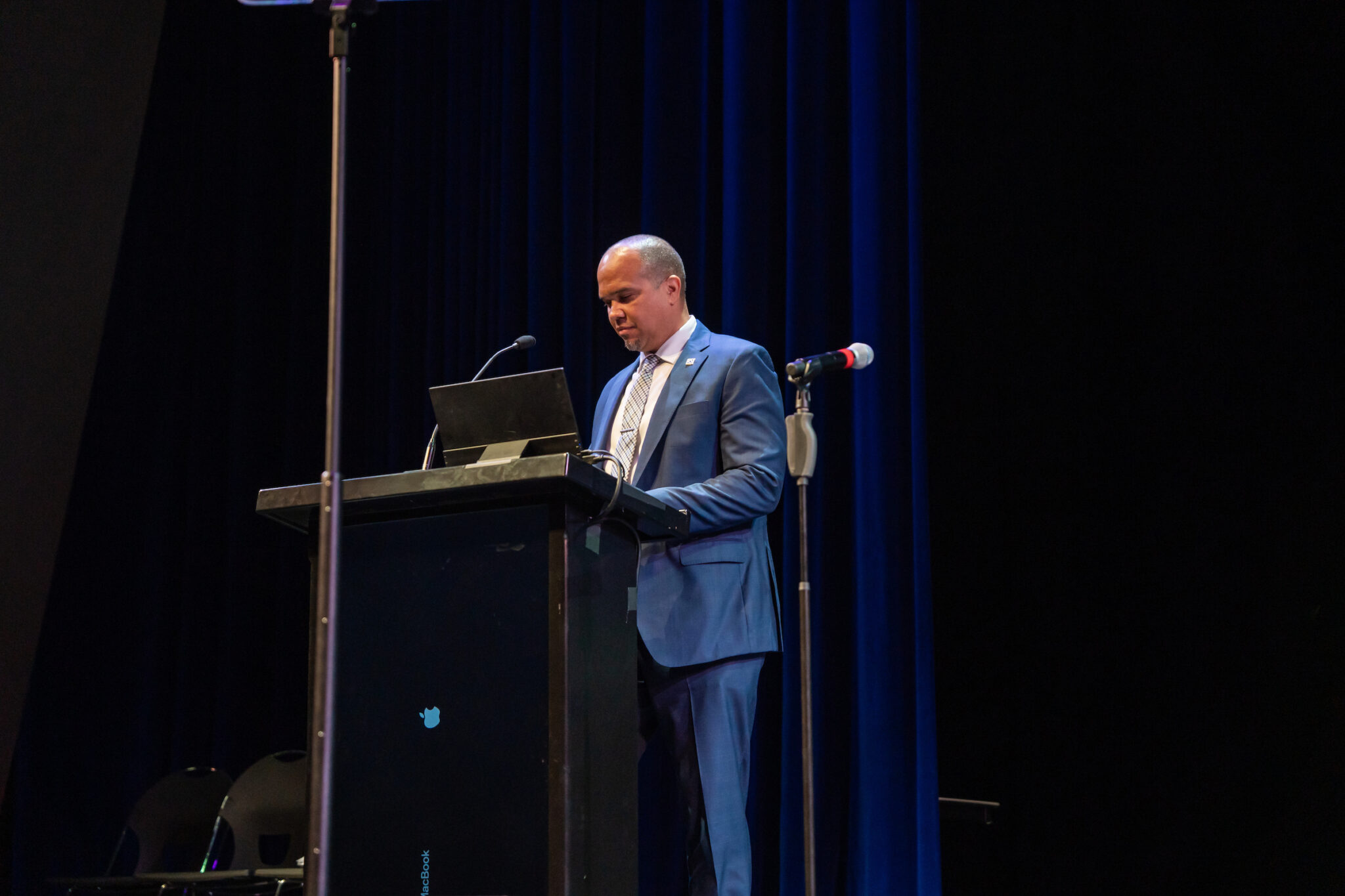 A person in a blue suit stands at a podium, looking down at a laptop while speaking into a microphone.