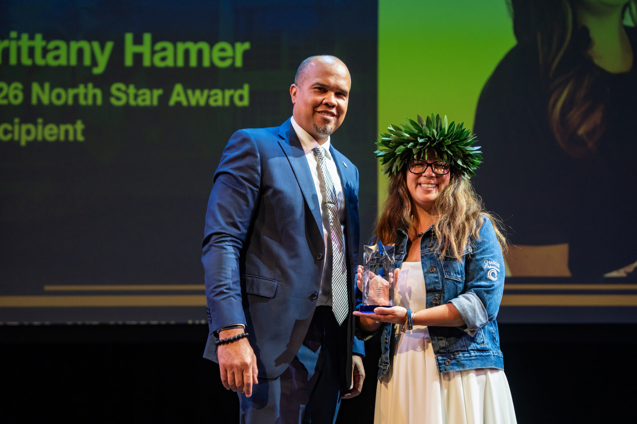 A person in a suit presents an award to another person wearing a leaf crown on stage,