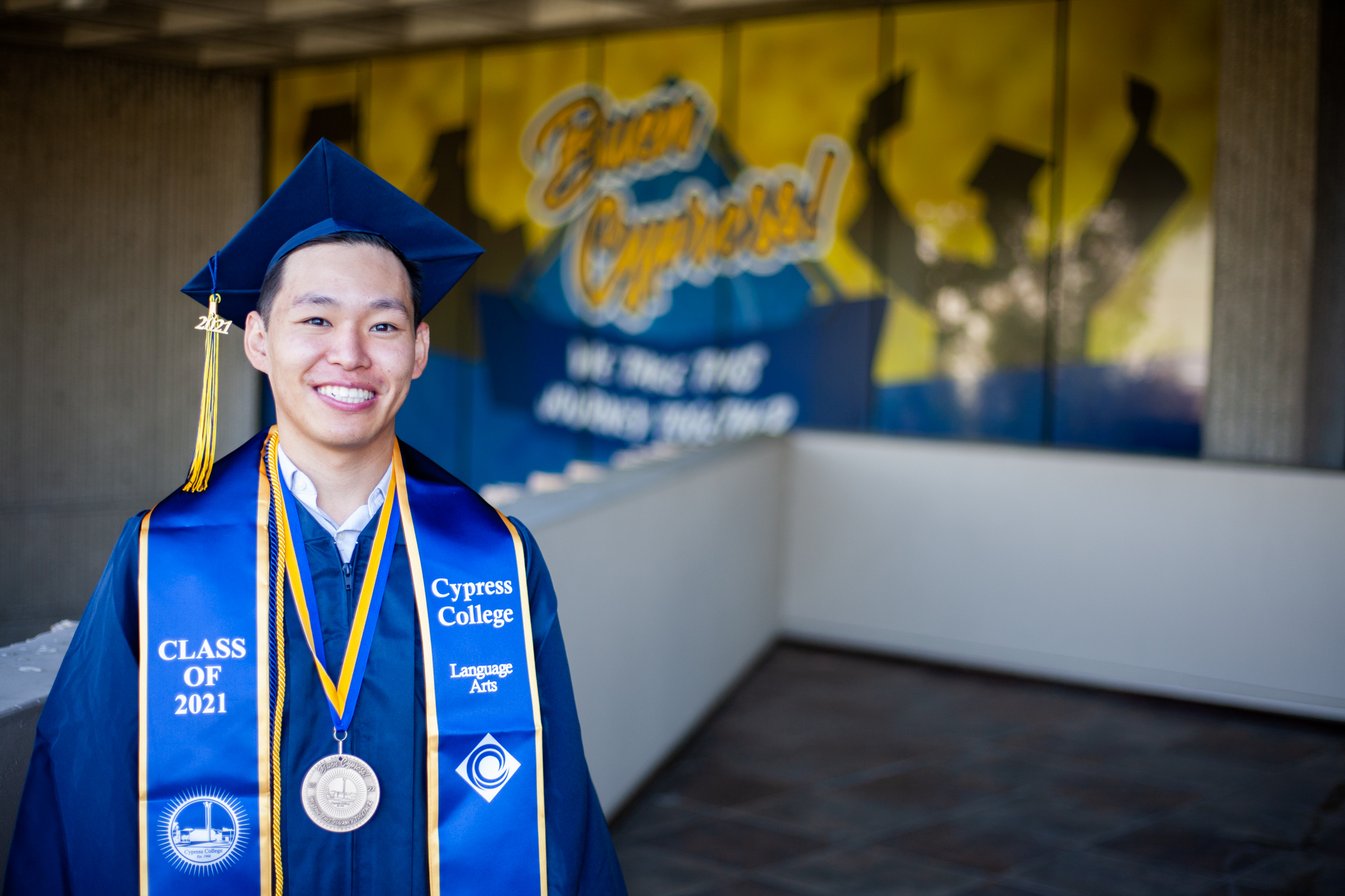 Student in graduation regalia poses in front of Buen Cypress backdrop.