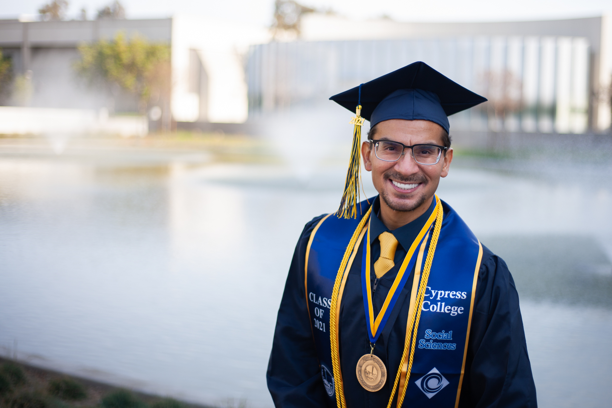 Student Wilfredo Carrasco wears graduation regalia and poses in front of Cypress College pond with fountain in background.
