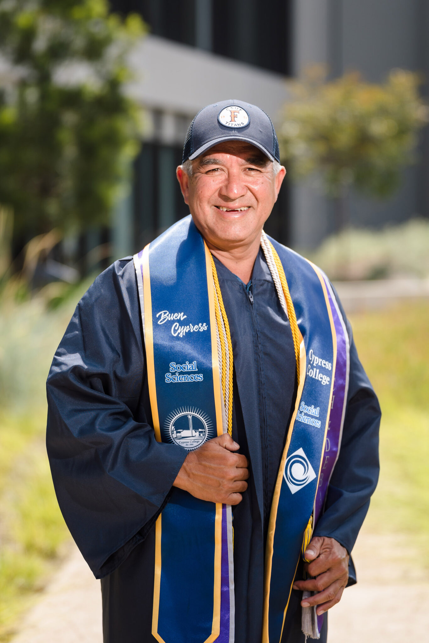 A gray-haired man in graduation regalia and a university baseball cap smiles at the camera. 
