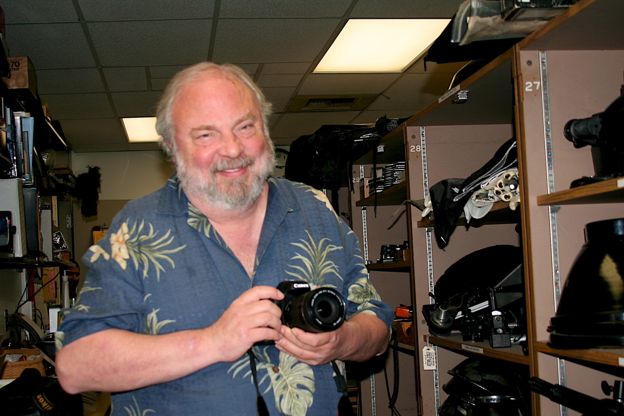 A smiling man with light hair and a beard holds a camera in a room with shelves of camera equipment.