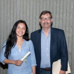 Young lady with long dark hair wearing a blue shirt, standing with a gentleman wearing glasses and a blue shirt and jacket.