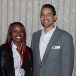 Young woman with red hair and black jacket smiling with a young man in glasses and a grey suit,