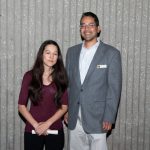 Young lady in a red top with long brown hair, standing with a young man in glasses and a grey suit,