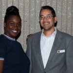 Young woman in a blue top with a young man in glasses and a grey suit.