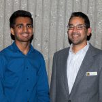 Man in a grey suit smiling standing with a young man wearing glasses in a blue shirt,