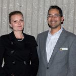 Young lady in a black outfit and white pearl necklace with a young man in glasses and a grey suit.