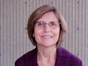 Older woman with short blond hair, smiling, wearing glasses and a maroon top
