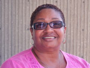 Black woman with short hair, smiling, wearing dark glasses, gold hoop earrings, and a pink top
