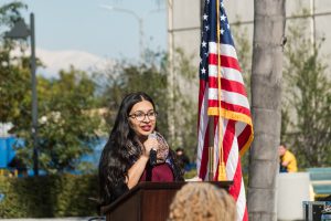 Woman with dark hair and glasses speaking at podium next to the American flag.