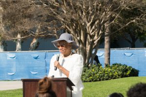 Woman with white dress, hat and sunglasses speaking at podium.