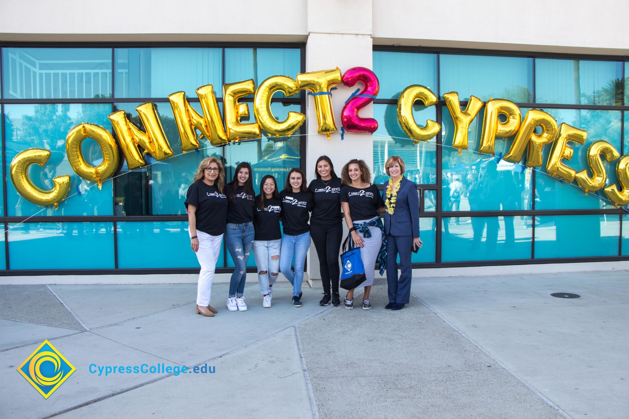 Dr. Schilling in a blue suit is standing with various staff who are all in black Connect2Cypress t shirts standing under gold and pink Connect2Cypress balloons.