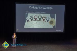 Dr. JoAnna Schilling in a brown pant suit, speaks on stage with a large projector with a graphic and the words "College Knowledge" behind her.