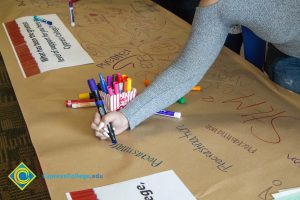 Someone writing "Procrastination" on a brown paper tablecloth with blue marker on a table covered in writing and a popcorn bucket full of colored markers.