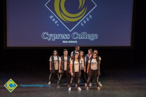 A group of students standing on stage in t shirts, sleeveless vests and pants with a large projector screen in the background.