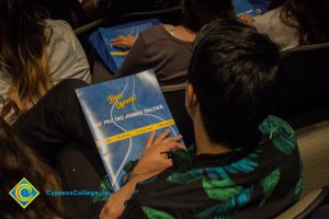 Students sitting in theater seats with blue handbooks on their lap.